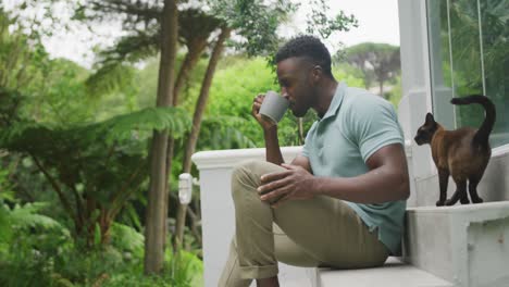 Happy-african-american-man-sitting-with-cat-and-drinking-coffee-in-garden