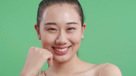 아름다운 아시아 여성이 한 손으로 얼굴을 만지고 미소 짓고 스튜디오에서 녹색 화면 배경에서 카메라를 보고 있습니다.