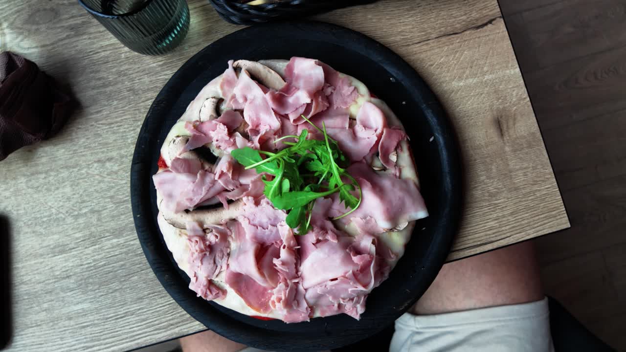Top view of a freshly served ham pizza with arugula on a black plate, placed on a wooden table, ready to eat. Perfect for culinary, dining, or lifestyle content