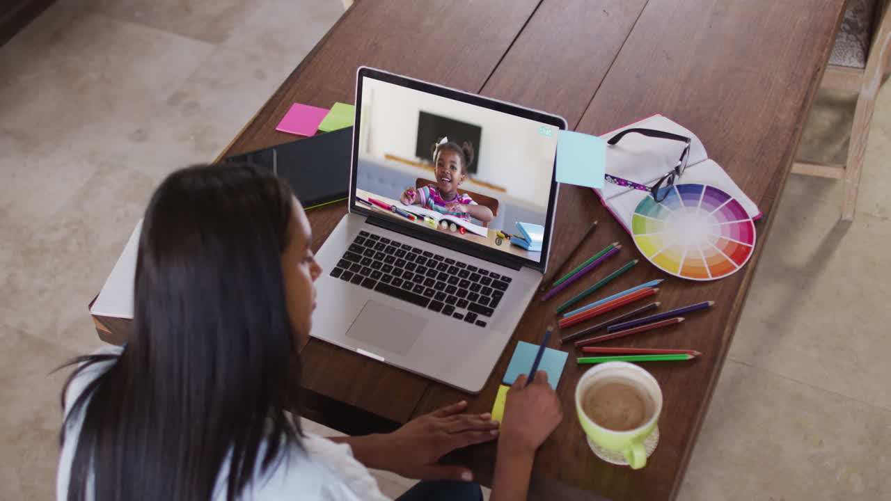 Maestra afroamericana escribiendo en notas adhesivas mientras realiza una videollamada con una niña en su computadora portátil.