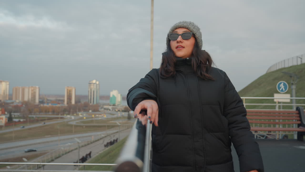 City bridge walk with dance rhythm, fullbody shot of woman in long coat strolling along metal walkway with skyline backdrop, cloudy sky, relaxed confident energy and cinematic urban panorama