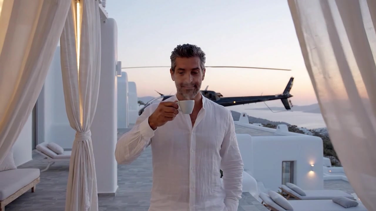 Uomo elegante in camicia bianca sorseggia caffè accanto a un elicottero di lusso al tramonto, godendosi una vista mozzafiato dalla terrazza di una villa che si affaccia su un paesaggio sereno.