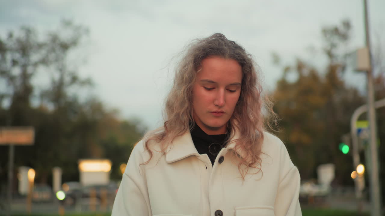Thoughtful girl in white coat walking alone with eyes closed along paved path surrounded by blurred view of urban background, distant cars, trees, and soft lights in calm peaceful evening atmosphere