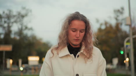 thoughtful girl in white coat walking alone with eyes closed along paved path surrounded by blurred view of urban background, distant cars, trees, and soft lights in calm peaceful evening atmosphere