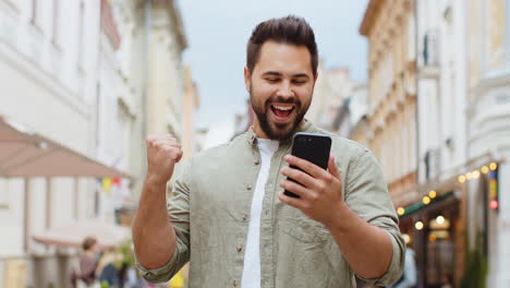 El-Hombre-Barbudo-Usa-Un-Teléfono-Inteligente-Móvil-Celebrando-Ganar-Buenas-Noticias-Al-Aire-Libre-En-Las-Calles-Urbanas-De-La-Ciudad