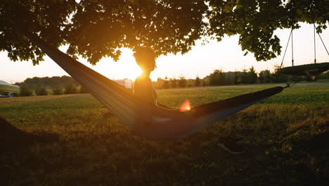 short-haired woman sits up in a hammock, looking out over a sunset casting a large lens flare