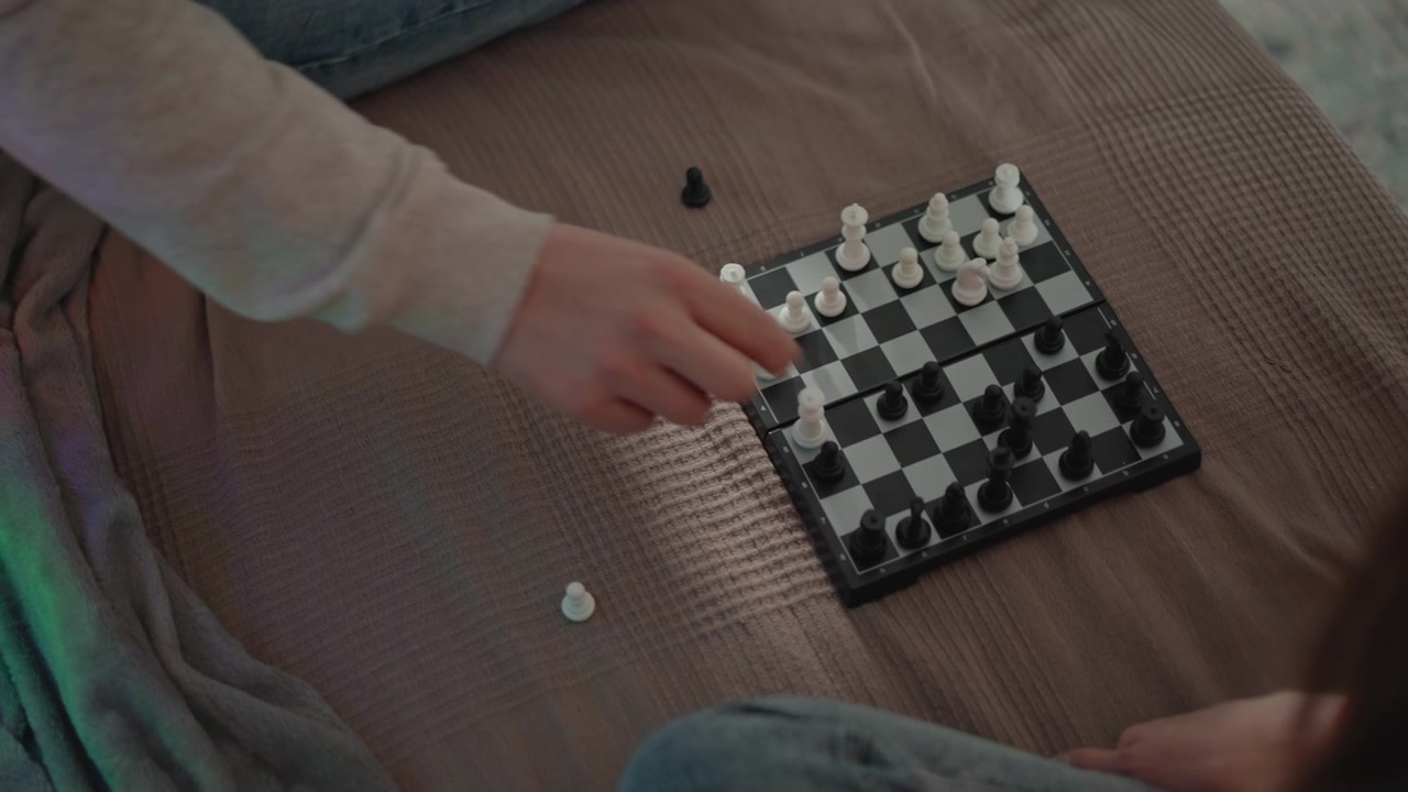 Due bambini seduti su un copriletto color tortora che giocano a scacchi su una piccola scacchiera, con una mano che si allunga per muovere o catturare un pezzo e pezzi caduti visibili accanto a un gioco rilassato al chiuso.