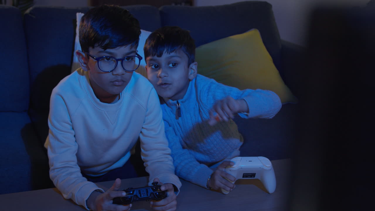 Two Young Boys At Home Playing With Computer Games Console On TV Holding Controllers Late At Night 4
