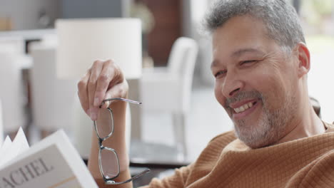 happy senior biracial man holding glasses, sitting reading book in living room, slow motion