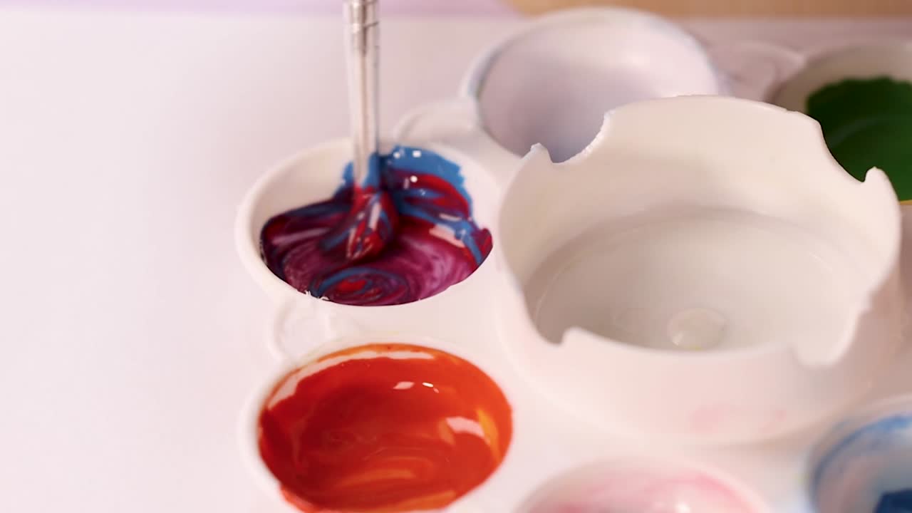 Un pennello mescola pigmenti rossi e blu in una palette bianca per creare un acquerello viola