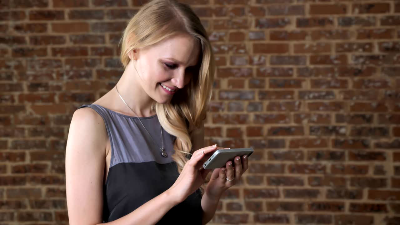 Ragazza giovane, bionda e carina guarda le foto sullo smartphone, sorridendo, concetto di comunicazione, sfondo di mattoni.