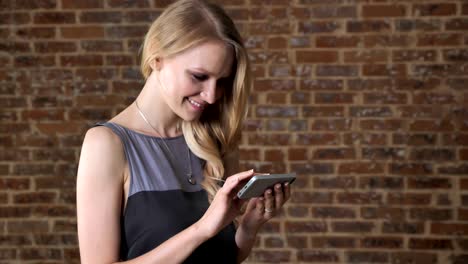 joven chica rubia linda está viendo fotos en el teléfono inteligente, sonriendo, concepto de comunicación, fondo de ladrillo