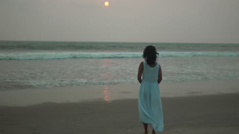 Una-Mujer-Vestida-De-Blanco-Observa-La-Puesta-De-Sol-En-Una-Playa-Serena,-Reflexionando-Sobre-La-Superficie-Del-Océano,-Un-Momento-Tranquilo