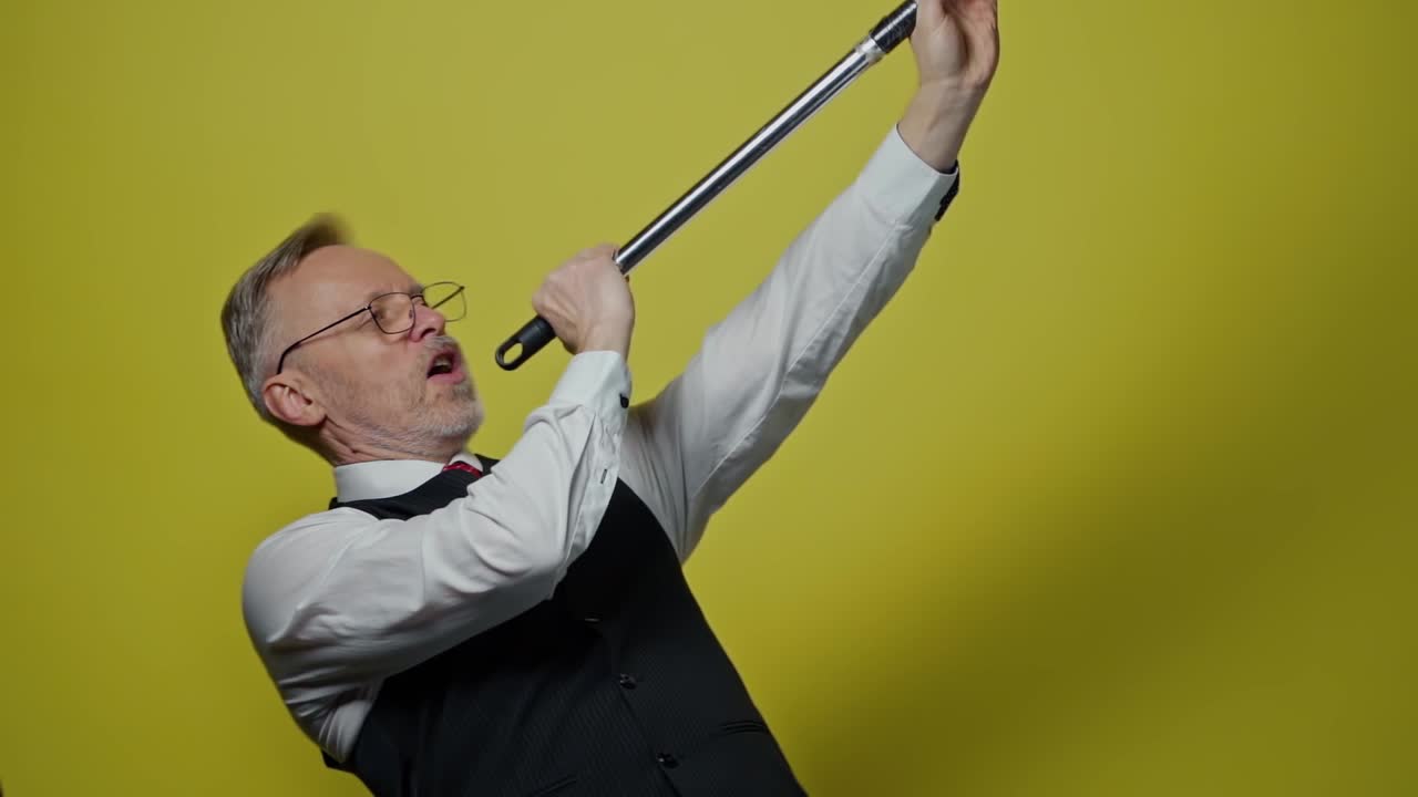 Mature businessman singing into the metal stick. Funny old man having joy with a mop like a microphone on the yellow studio background. Slow motion.