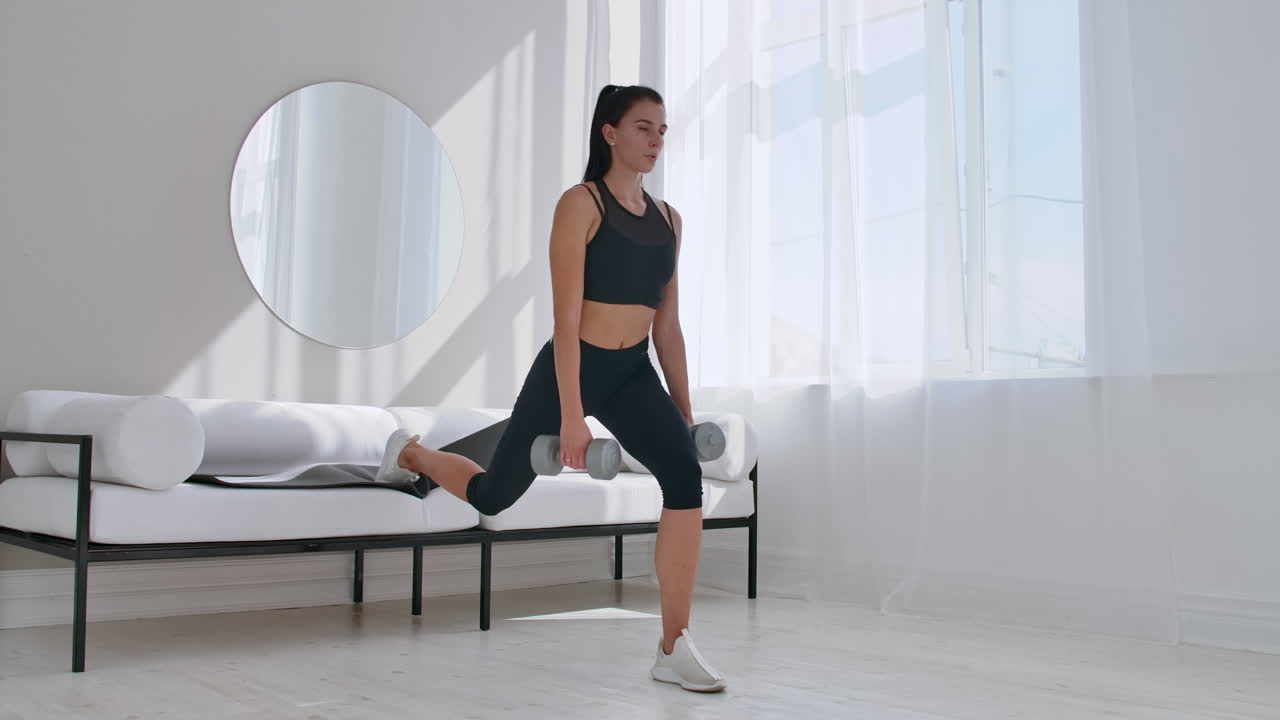 Una bruna in abbigliamento sportivo nero in un appartamento bianco esegue uno split squat con manubri tra le mani, appoggiando il piede sul divano.