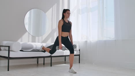 brunette in black sportswear in a white apartment makes a split squat with dumbbells in her hands leaning on the sofa with her foot.