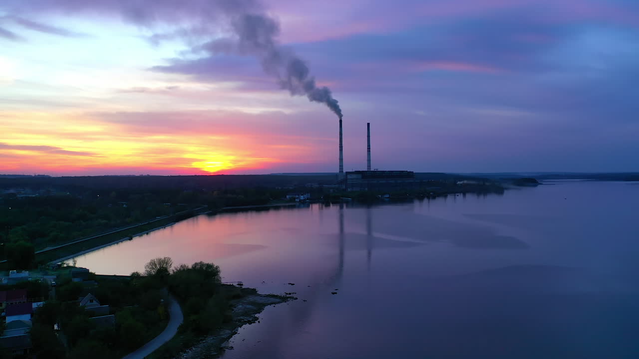 Pipes with smoke on beautiful nature background. Harmful manufacturing near the river in the evening. Blue color of water and sky near industry at sunset. Aerial view.