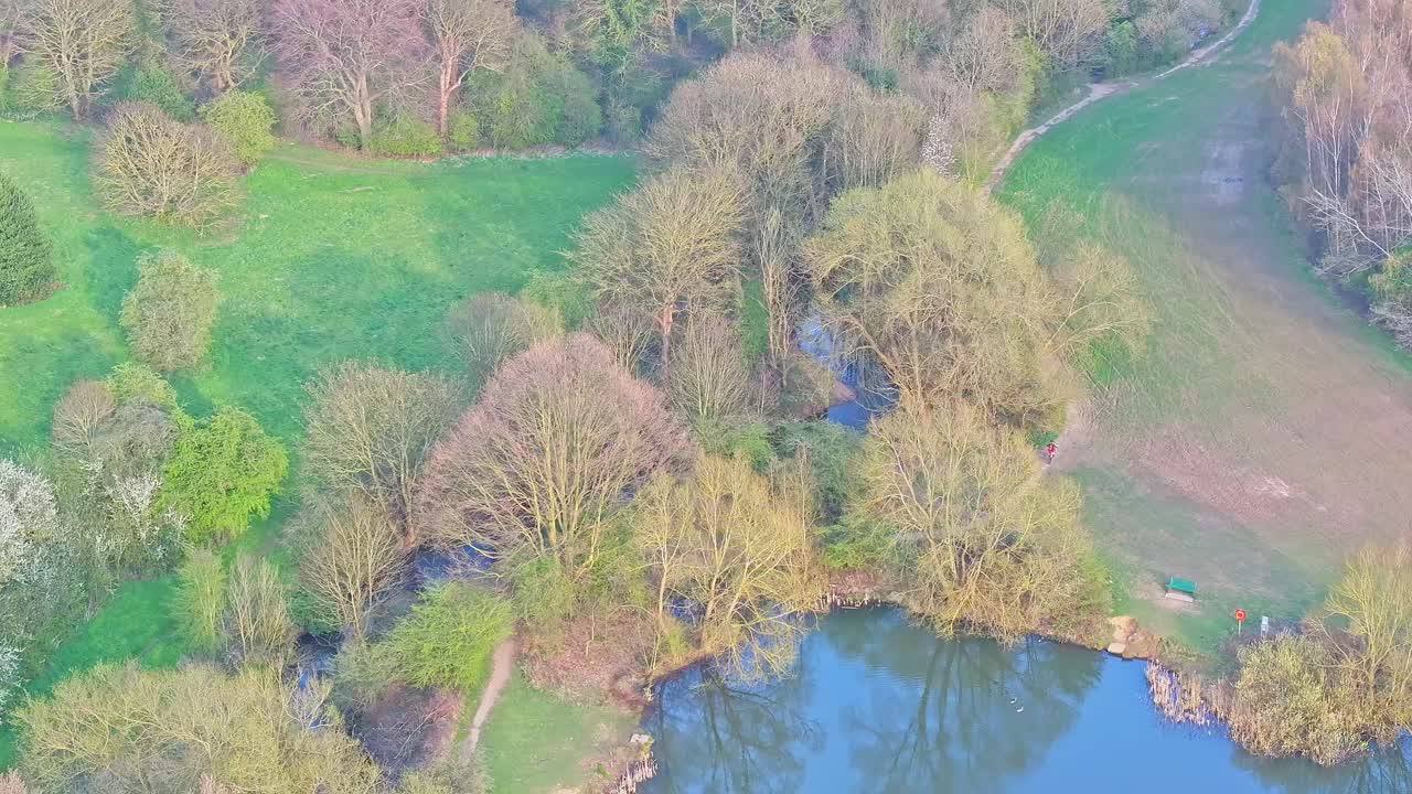 Drone aerial showing a reflective pond bordered by grassy banks and tree-lined edges, walking paths, and early spring foliage in Netherwood Country Park, Wombwell, South Yorkshire