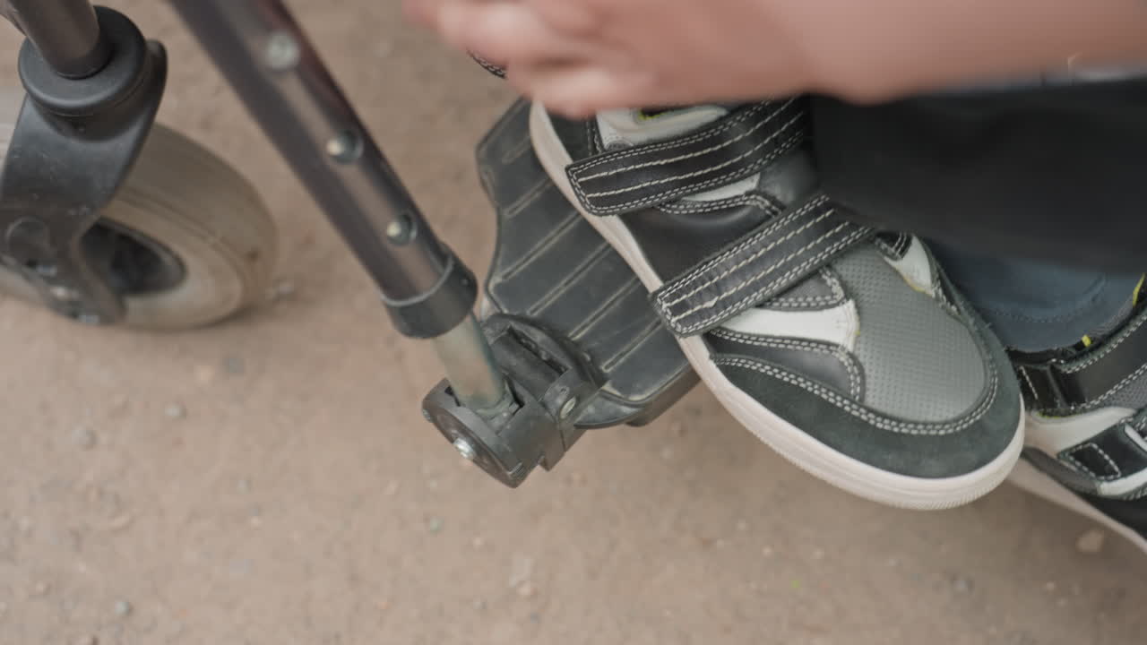 CloseUp Of Hand Adjusting Footrest Strap Over Sneaker, Velcro And Buckle Action, Careful Placement For Safe Travel, Visible Wheel And Dusty Ground In Outdoor Setting Showing Preparation