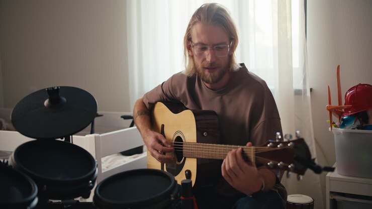 A blond man in glasses with a beard in a brown T-shirt plays a yellow acoustic guitar near him there is a drum set development of talents and hobbies at home