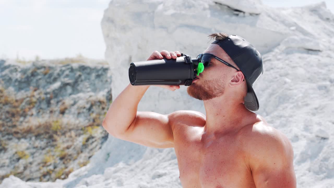 A strong naked man opens a black bottle and drinks water. Outdoor fitness