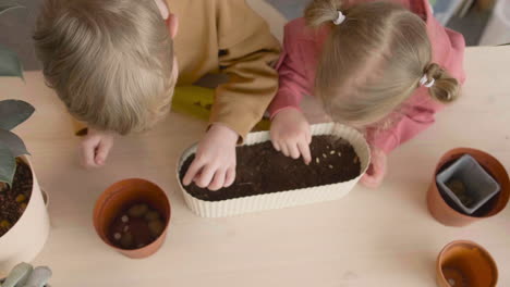 Vista-Superior-De-Una-Niña-Rubia-Y-Un-Niño-Rubio-Preparando-El-Suelo-En-Una-Olla-Sentada-En-Una-Mesa-Donde-Hay-Plantas-En-Un-Taller-De-Artesanía