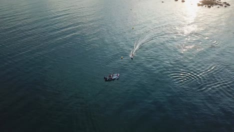 Overhead-aerial-shot-of-surfer-riding-electric-board-past-small-boat-with-golden-hour-light-reflection-on-water-surface