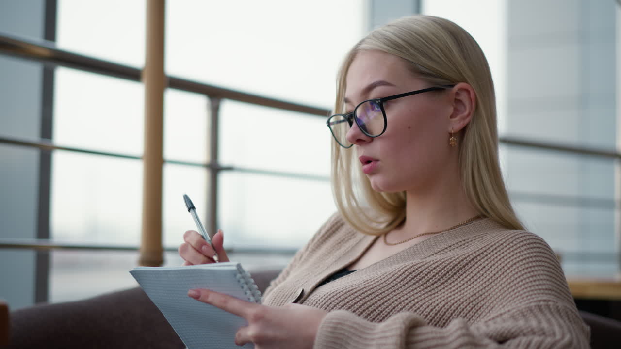 Young woman thoughtfully taking notes in her notebook, she is focused and deep in thought, writing in a quiet setting, possibly a cafe or study area