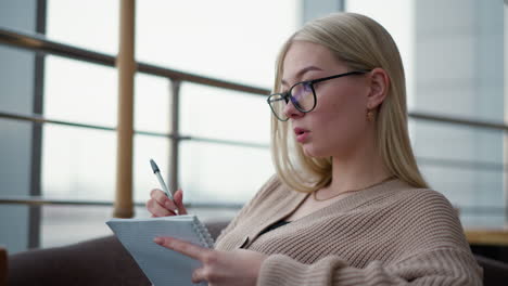 mujer joven pensativa tomando notas en su cuaderno, está enfocada y profundamente pensativa, escribiendo en un entorno tranquilo, posiblemente una cafetería o área de estudio
