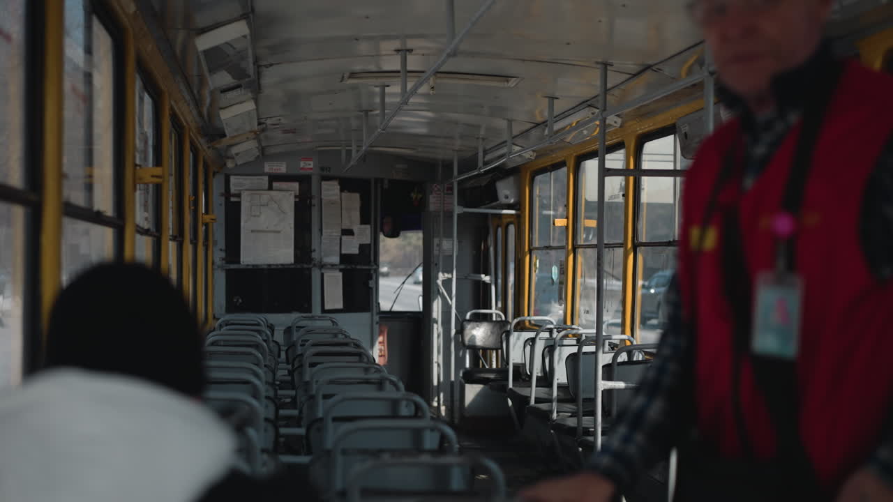 near empty winter train interior shows rows of metal seats, one passenger seated while other rider stands with tag on neck, windows reveal snowy city street outside, quiet motion