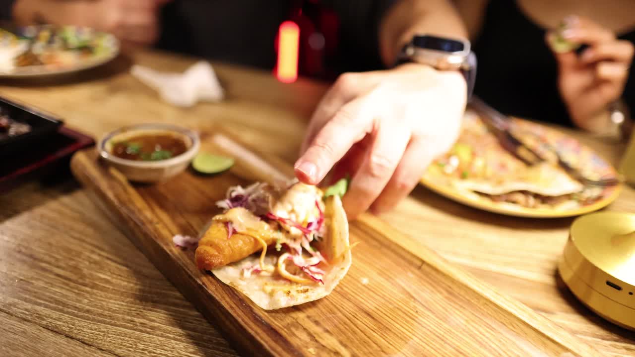 A person assembles tacos on a wooden board under warm lighting in a cozy restaurant setting