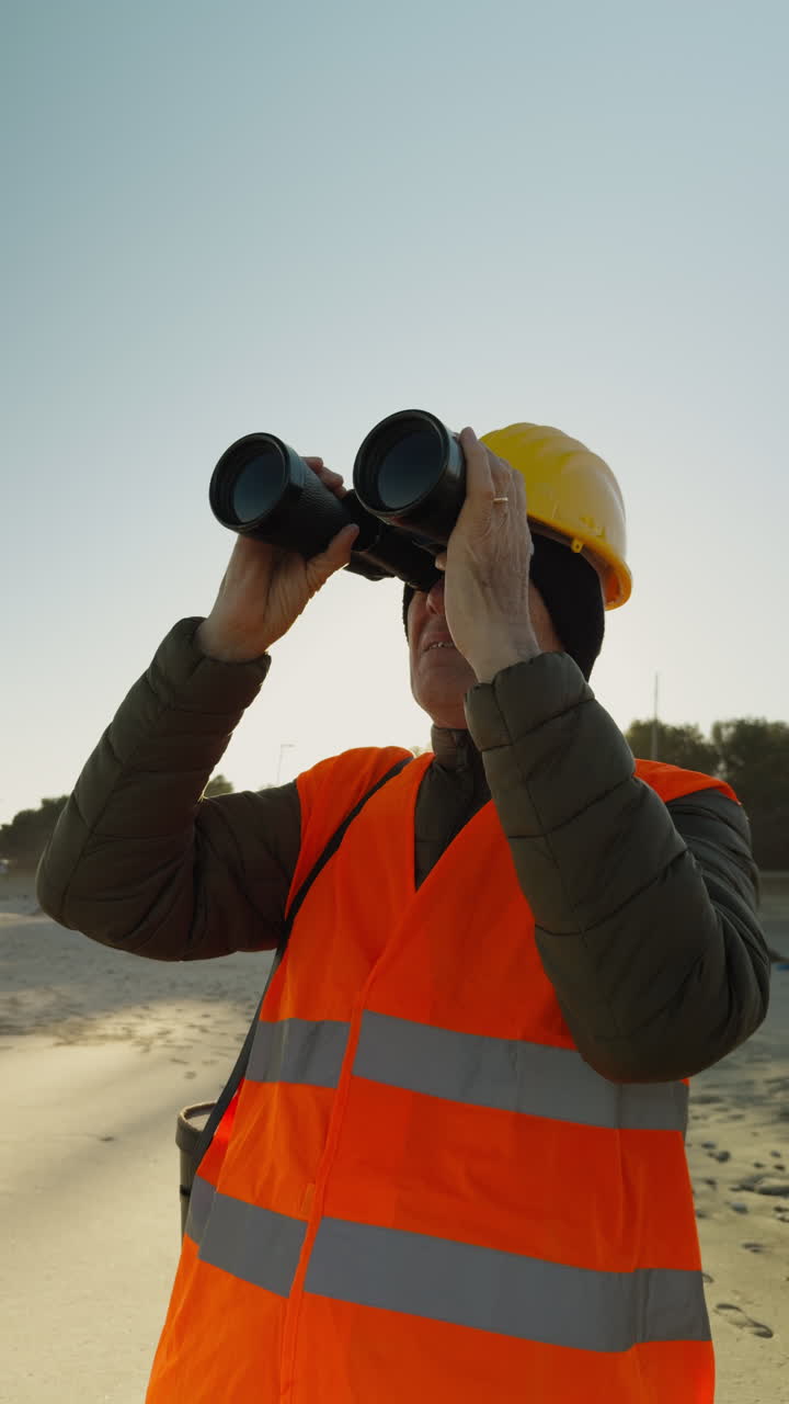 En arbejder iført refleksvest og hjelm ser ud over havet gennem en kikkert