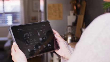 video of midsection of biracial woman at home holding tablet with smart home interface screen