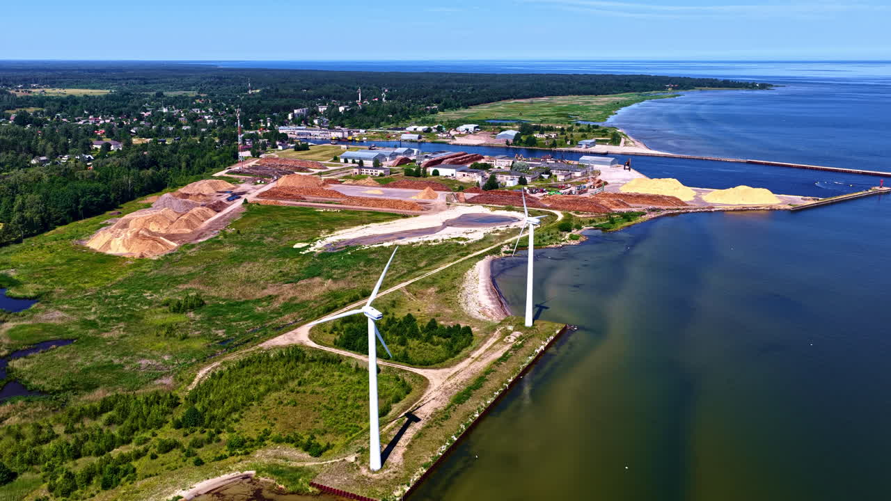 Aerial view of Engure, Latvia, showing wind turbines, timber storage, and the coastline along the Baltic Sea on a clear summer day