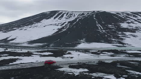 Conducir-En-Islandia-En-La-Temporada-De-Primavera,-Vista-Aérea-De-Un-Coche-Rojo-En-Una-Carretera-Mojada-Bajo-Colinas-Volcánicas-Cubiertas-De-Nieve