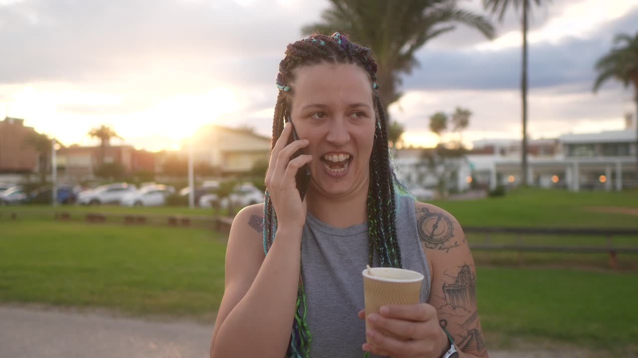 mujer con trenzas y sonrisa hablando en un teléfono inteligente con café en la mano