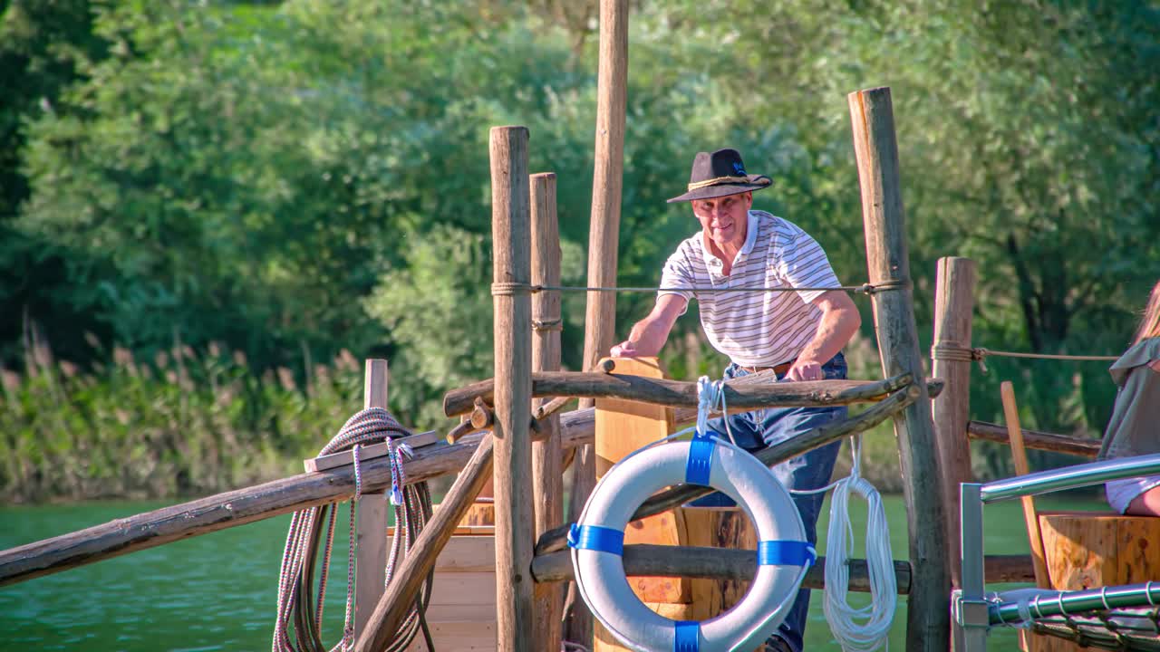 Elderly man greets camera from a moving boat in green landscape. Tracking shot