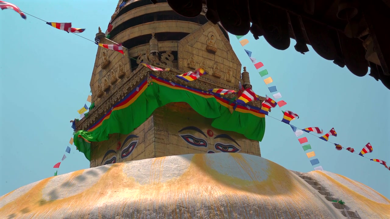 Spårningsbild tagen från låg vinkel av Swayambhunath Stupas gyllene spira och ikoniska visdomsögon, dekorerad med färgglada buddhistiska flaggor som vajar under en blå himmel i Katmandu, Nepal.