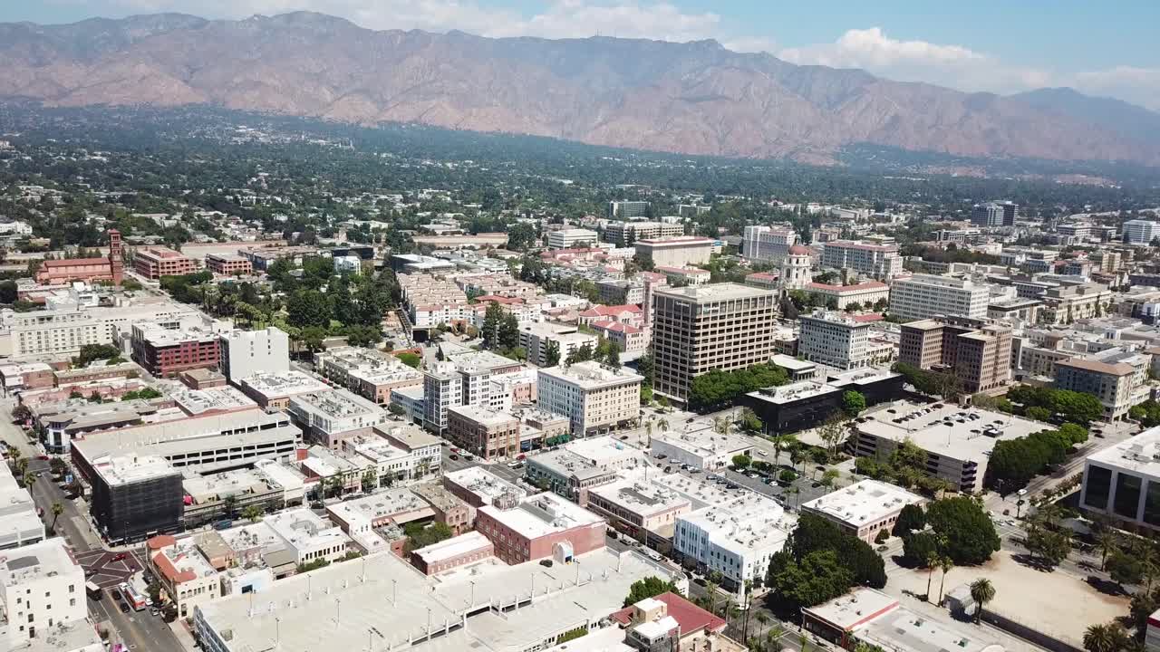 Ampia ripresa aerea che si allontana dal centro di Pasadena, in California. La telecamera si ritrae per rivelare una vista più ampia di isolati di media altezza, strade alberate e le montagne di San Gabriel
