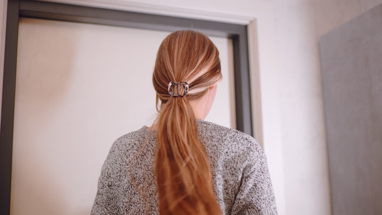 Side profile of woman closing door after preparing to leave home, wearing gray sweater and jeans, holding black jacket in hand, hair tied with clip, step outside with confident expression