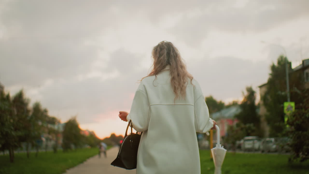 back view of woman walking outdoors carrying handbag and twirling white umbrella with curly hair flowing freely while cars pass by and people move in blurred background