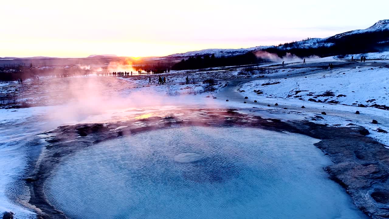 панорамный вид с дроном гейзера в гейзире, исландия. дым выходит из гейзера с людьми, гуляющими вокруг гейзера, под красным и оранжевым закатом.