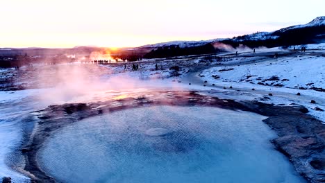 panoramic view with a drone of a geyser in geysir, iceland. smoke is getting out of the geyser with people walking all around the geyser, under the red and orange sunset.