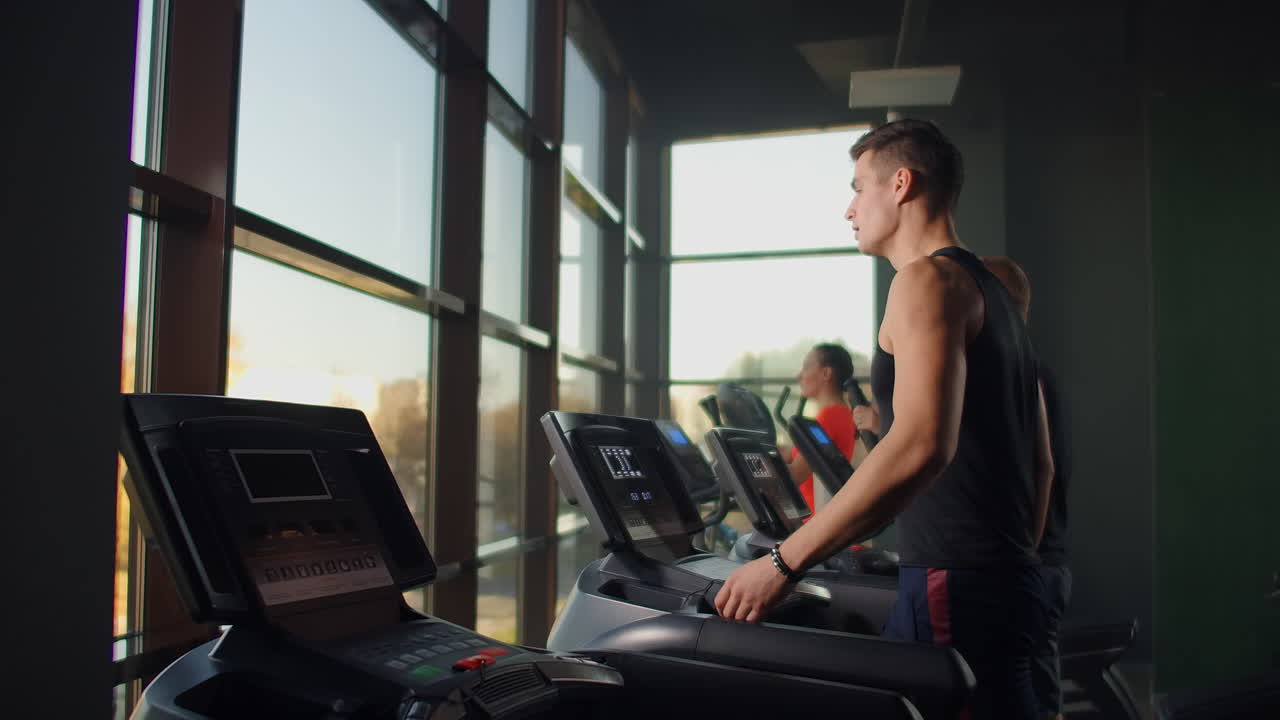 Men walk on treadmills in the fitness room. A group of people walking on treadmills near a large panoramic window. Start of fitness classes. Young beautiful women and men in sportswear in cardio fitness room