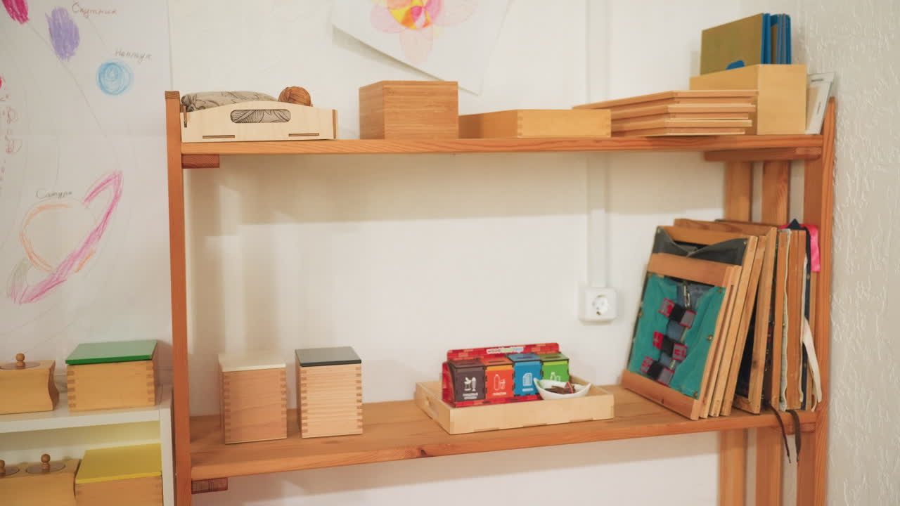 Wooden shelf displays variety of Montessori-inspired educational toys and learning tools including dressing frames, storage boxes, and fabric panels arranged neatly in bright classroom