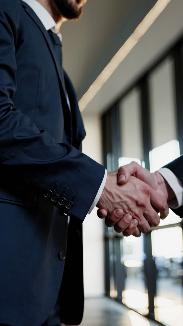 Low-angle video shot of two people in suits shaking hands in a modern office, symbolizing business