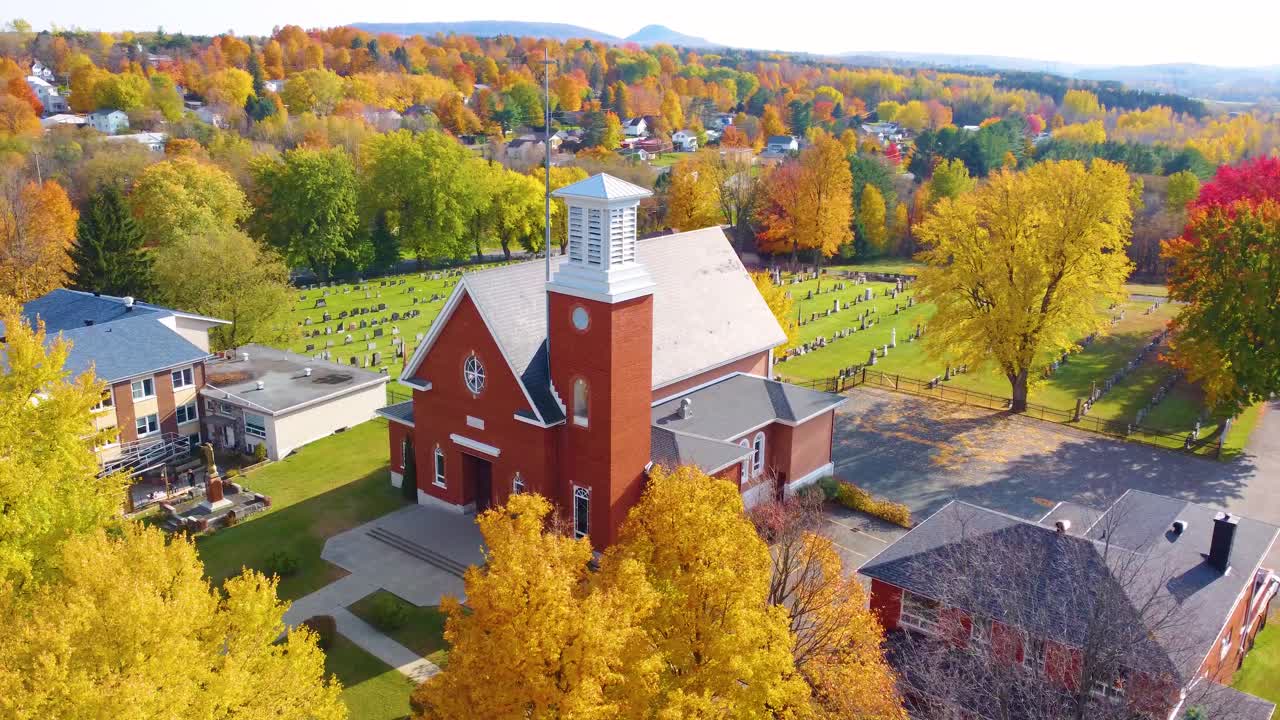 Aerial view of a red brick church near a cemetery surrounded by colorful trees during fall season in the Estrie region of Quebec, Canada. Orbit Motion Shot