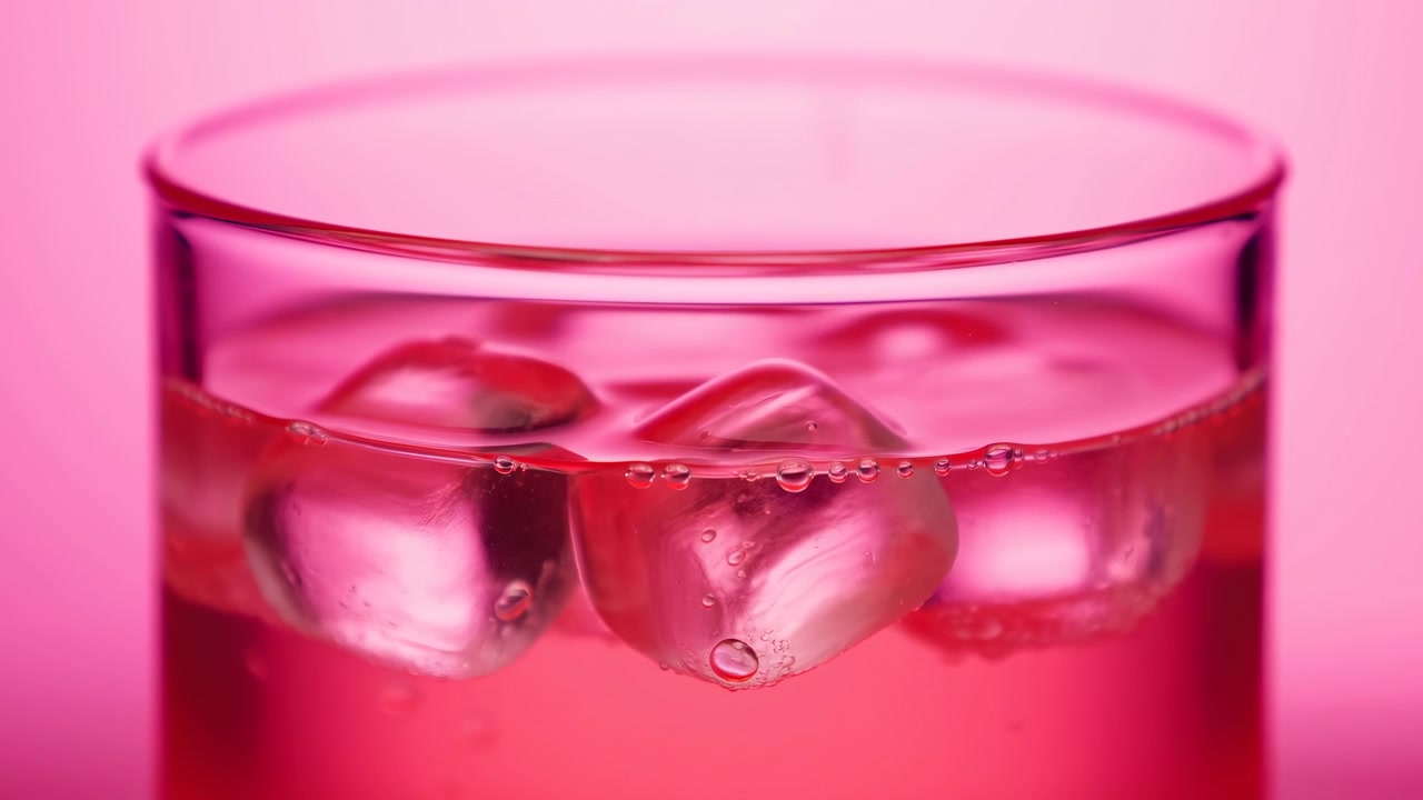 Vista a la altura de los ojos de un vaso cilíndrico transparente parcialmente lleno de una bebida translúcida de color rosa rojizo y cubitos de hielo en la superficie, con formas flotantes, burbujas, reflejos húmedos y un fondo de estudio limpio para.