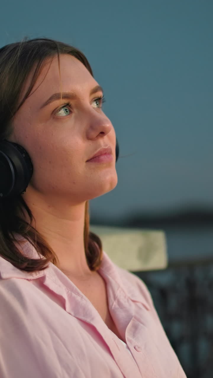 joven con camiseta rosa y auriculares descansando en la valla, mirando contemplativamente a la luna en el fondo, pequeño insecto negro volando cerca de su cabeza, con un entorno al aire libre pacífico