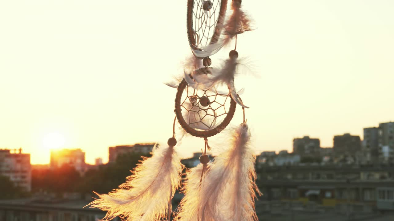 amuleto sueña catcher agitando en un viento ligero contra el telón de fondo de una puesta de sol brillante y un bokeh de luces de la ciudad. plumas de luz blanca se elevan en el viento. el disco del sol poniente. amuleto étnico